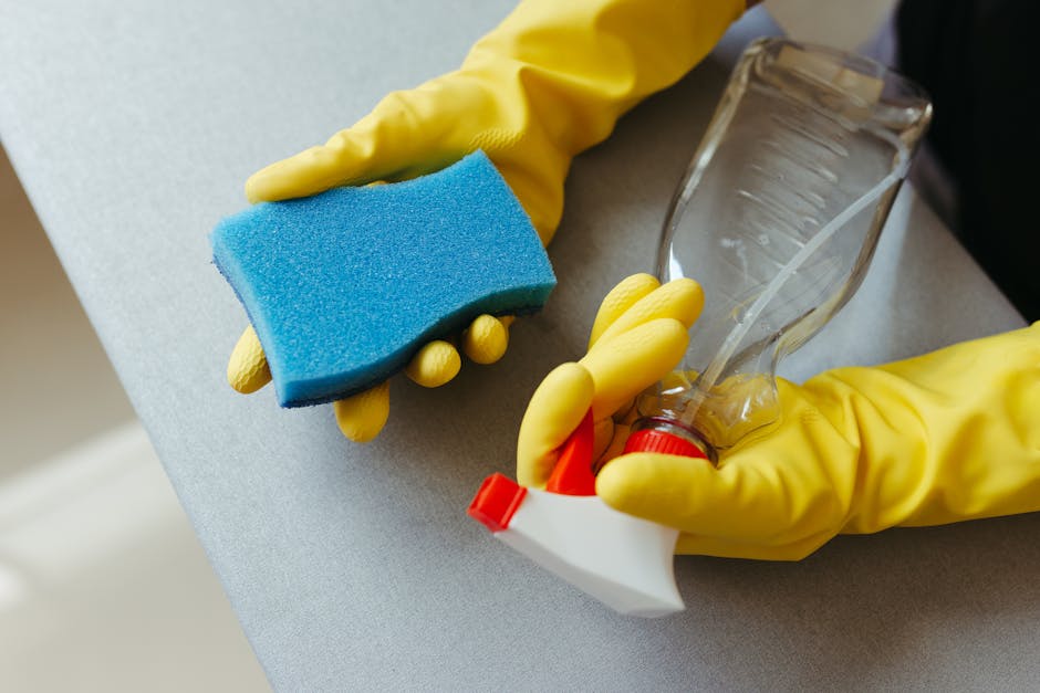 A close-up view of a person wearing yellow rubber gloves holding a blue scrub sponge in their right hand and a spray bottle with a clear nozzle in their left hand. The background shows a light-colored, smooth countertop surface, likely in a domestic or commercial cleaning setting. The environment appears clean and well-maintained, with the focus on cleaning tools used for surface cleaning or sanitisation. The scene emphasizes the cleaning process, aligned with professional efforts by Carpet Cleaning SE11, as seen on the page about avoiding common cleaning mistakes in Kennington flats, SE11.