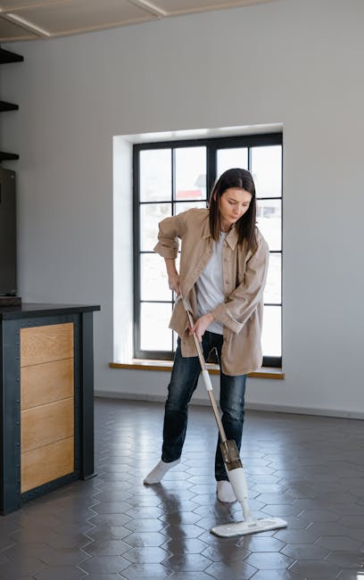 A woman engaged in domestic cleaning stands on a dark hexagonal tiled floor in a modern, well-lit room with a large window featuring black grid framing. She is using a mop to clean the surface, appearing focused on maintaining hygiene and cleanliness. The room has white-painted walls and minimalistic furniture, including a wooden cabinet with black fittings visible on the left side. Natural light floods the space through the window, illuminating the clean, shiny floor. This scene reflects routine surface cleaning and deep cleaning practices, highlighting the importance of proper sanitisation in residential environments. The professional cleaning service, Carpet Cleaning SE11, provides expert assistance with keeping flats in Kennington SE11 spotless and hygienic, emphasizing high standards of domestic cleaning.