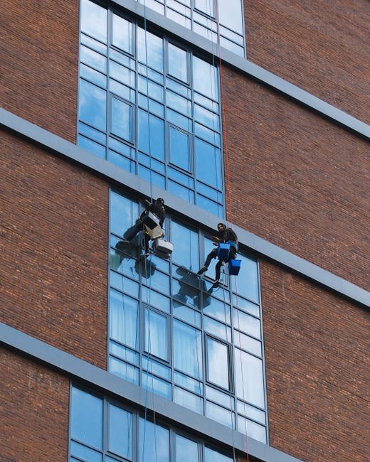 A window cleaner suspended by ropes and harnesses is performing high-level surface cleaning on a modern glass facade in an urban building. The window cleaner is wearing safety gear, including a helmet