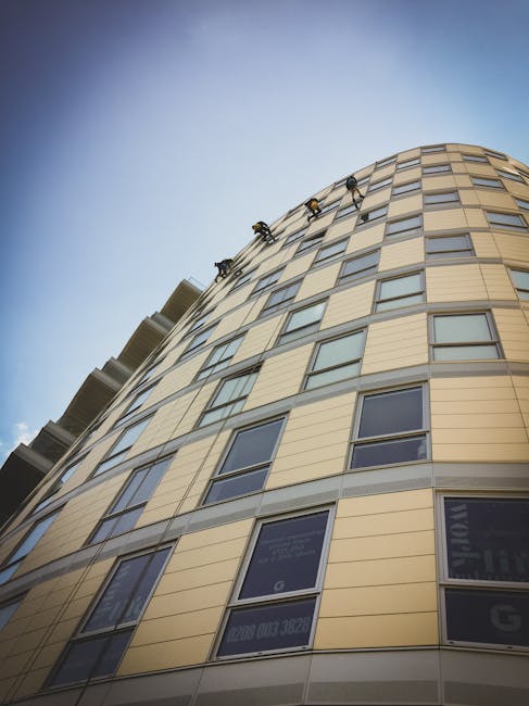 The image shows a modern, curved high-rise residential building with a yellow and beige exterior, featuring multiple rows of large rectangular windows. The building's surface appears clean and well-maintained, with some windows reflecting the blue sky and sunlight. There are two window cleaners visible on the upper part of the building, suspended by ropes and performing surface cleaning tasks on the glass and facade panels. The scene is illuminated by natural daylight, highlighting the building's sleek architectural design. This setting emphasizes the importance of professional cleaning services for maintaining the appearance and hygiene of commercial or residential properties, aligning with the focus on end of tenancy cleaning for SE11 flats on Kennington Road provided by Carpet Cleaning SE11.
