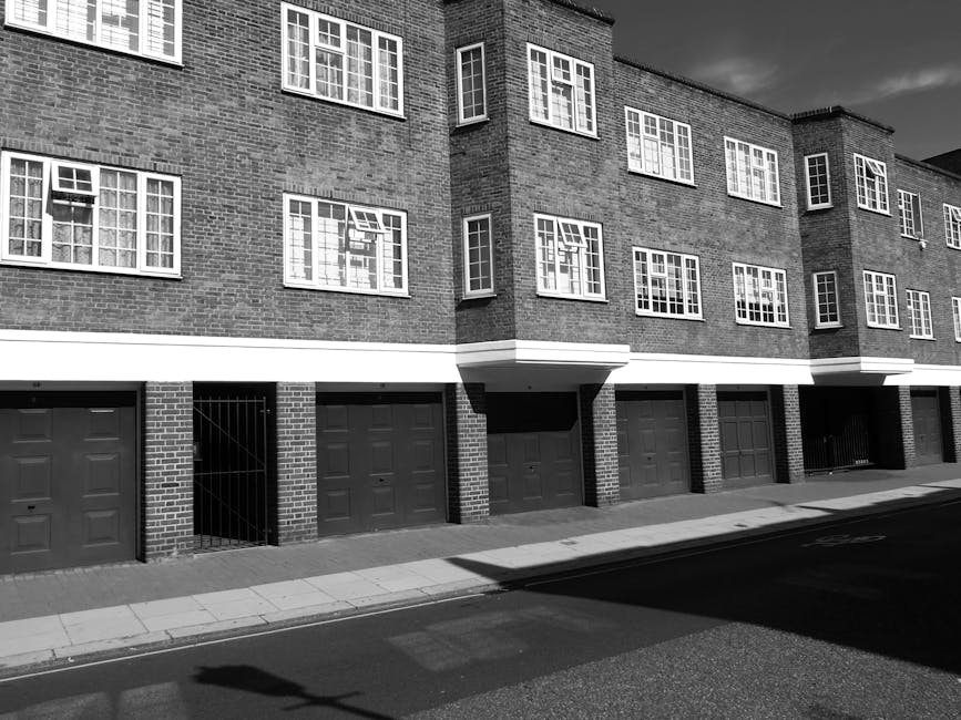 Black and white photograph of a modern residential building with multiple garage doors at street level, constructed with brick and featuring large, evenly spaced windows on the upper floors. The surface of the street is paved with asphalt, and the sidewalk is made of concrete slabs. The building's exterior appears clean and well-maintained, with shadows cast by sunlight highlighting the architectural details. The overall scene depicts an urban housing complex in the vicinity of Kennington Road, suggesting a tidy and orderly environment, consistent with the cleaning and maintenance services offered by Carpet Cleaning SE11 for residential properties.