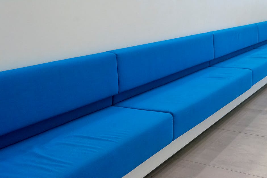 A long, modern seating bench upholstered in bright blue fabric, situated against a plain white wall in an indoor setting, with smooth, clean surface and visible cushion seams. The floor beneath is light-colored and appears well-maintained, reflecting a tidy appearance. No additional objects or tools are visible, indicating a neat and hygienic environment. This image exemplifies surface cleaning and maintenance, aligning with professional domestic or commercial cleaning standards provided by Carpet Cleaning SE11, situated in a well-lit, orderly space suitable for hygienic and deep cleaning services in the Kennington area, SE11.