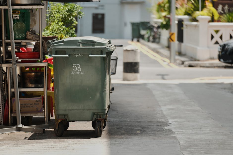 A large green outdoor wheeled trash bin positioned on the sidewalk near a residential building, with visible wheels and a closed lid. To the left of the bin, there is a metal shelving unit with cleani