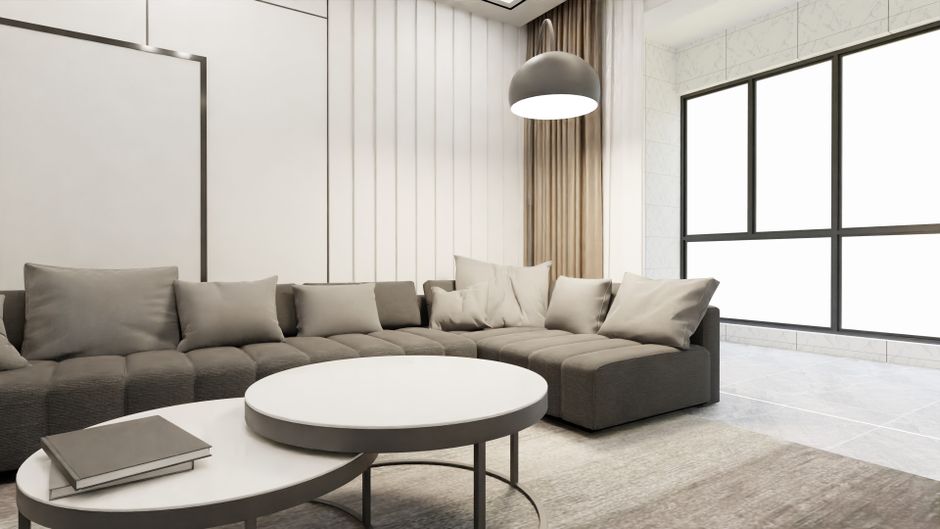 A modern living room featuring a large L-shaped grey fabric sofa with several matching cushions, positioned against a white-paneled wall. In front of the sofa, there are two overlapping round coffee t