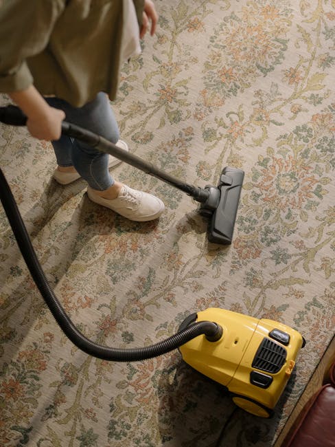 A person operating a yellow vacuum cleaner with a black hose, cleaning a large, patterned area rug with floral motifs in shades of beige, green, and orange. The person is dressed in casual attire, including white sneakers and light-colored pants, standing on the rug in a room with wooden flooring visible at the edges. The lighting appears natural and even, highlighting the clean, dust-free surface of the rug, which is being deep cleaned as part of a professional domestic or commercial surface cleaning service. The scene emphasizes thorough hygiene and maintenance, aligning with the services provided by Carpet Cleaning SE11 for rugs near Kennington Oval and Vauxhall, SE11.