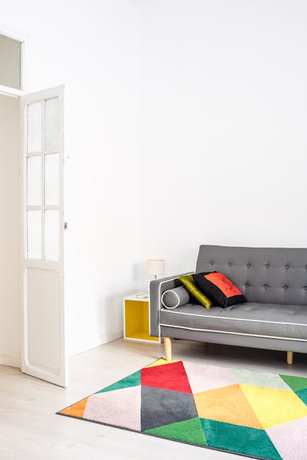 A modern living room featuring a light-colored hardwood floor and a multicolored geometric area rug. The room has white walls and an open white door with glass panels on the left, allowing natural light to brighten the space. A grey upholstered sofa with wooden legs is positioned against the right wall, decorated with three cushions in black, yellow, and red. Next to the sofa, there is a small yellow side table with a white table lamp. The surfaces and flooring appear clean and free of dust or stains, highlighting the professional cleaning and maintenance provided by Carpet Cleaning SE11 for domestic environments, specifically in the context of surface cleaning and sanitation near Kennington Oval and Vauxhall, SE11.