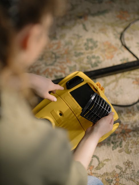 A person holding a yellow portable carpet cleaning device with a black vent, preparing for surface cleaning or deep cleaning in a room with a patterned carpet featuring shades of beige, orange, and green. The individual’s hands are visible, and they are seated or kneeling on the carpet, with the device positioned close to the floor. The background shows natural lighting, highlighting the cleanliness of the surface. The setting emphasizes professional domestic or commercial carpet sanitisation, as promoted by Carpet Cleaning SE11, and reflects a focus on maintaining hygiene and clean surfaces in residential or commercial spaces within the SE11 area.