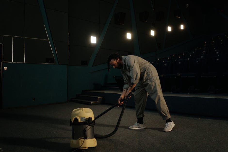 A person holding a yellow portable carpet cleaning device with a black vent, preparing for surface cleaning or deep cleaning in a room with a patterned carpet featuring shades of beige, orange, and green. The individual’s hands are visible, and they are seated or kneeling on the carpet, with the device positioned close to the floor. The background shows natural lighting, highlighting the cleanliness of the surface. The setting emphasizes professional domestic or commercial carpet sanitisation, as promoted by Carpet Cleaning SE11, and reflects a focus on maintaining hygiene and clean surfaces in residential or commercial spaces within the SE11 area.