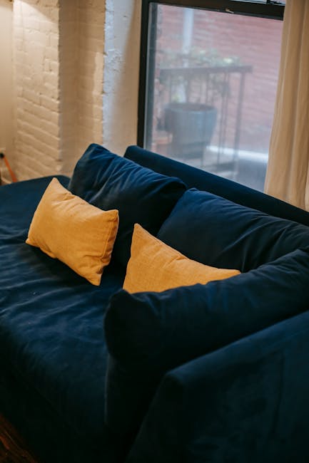 Close-up view of a dark blue fabric sofa with two mustard-yellow cushions placed on it, situated near a large window that allows natural light to brighten the room. The window reveals an outdoor balcony with potted plants and a red brick wall in the background. The sofa appears clean and well-maintained, with no visible stains or dust, highlighting the surface cleaning and maintenance typical of domestic cleaning services provided by Carpet Cleaning SE11. The scene emphasizes a tidy, inviting living space, suitable for deep cleaning and sanitisation to ensure hygiene and comfort in a residential setting.