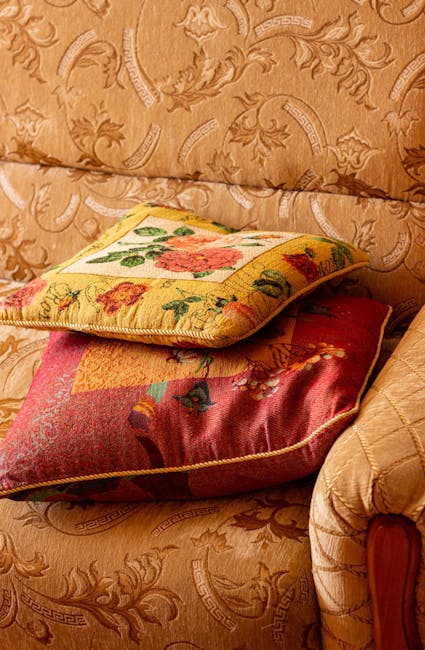 Close-up of two decorative cushions placed on a patterned fabric sofa in a living room. One cushion features a floral tapestry design with a yellow background, while the other has a multicolored, textured fabric with a red base. The sofa's fabric has an intricate, regal pattern with a subtle sheen, suggesting a smooth, soft material. The scene is illuminated by warm ambient lighting, highlighting the cleanliness and well-maintained appearance of the upholstery. This setting exemplifies domestic surface cleaning and maintenance, aligning with Carpet Cleaning SE11's services for sofa cleaning for Kennington Lane residents, SE11.